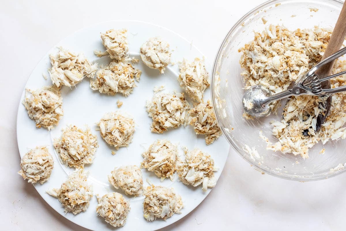 A plate with clusters of crab ball mixture sits next to a glass bowl containing more of the same mixture and a metal scoop.