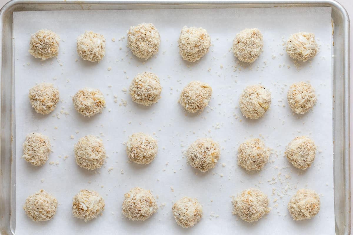 A baking sheet lined with parchment paper holds twenty four evenly spaced, round, breaded crab balls, ready to be baked.