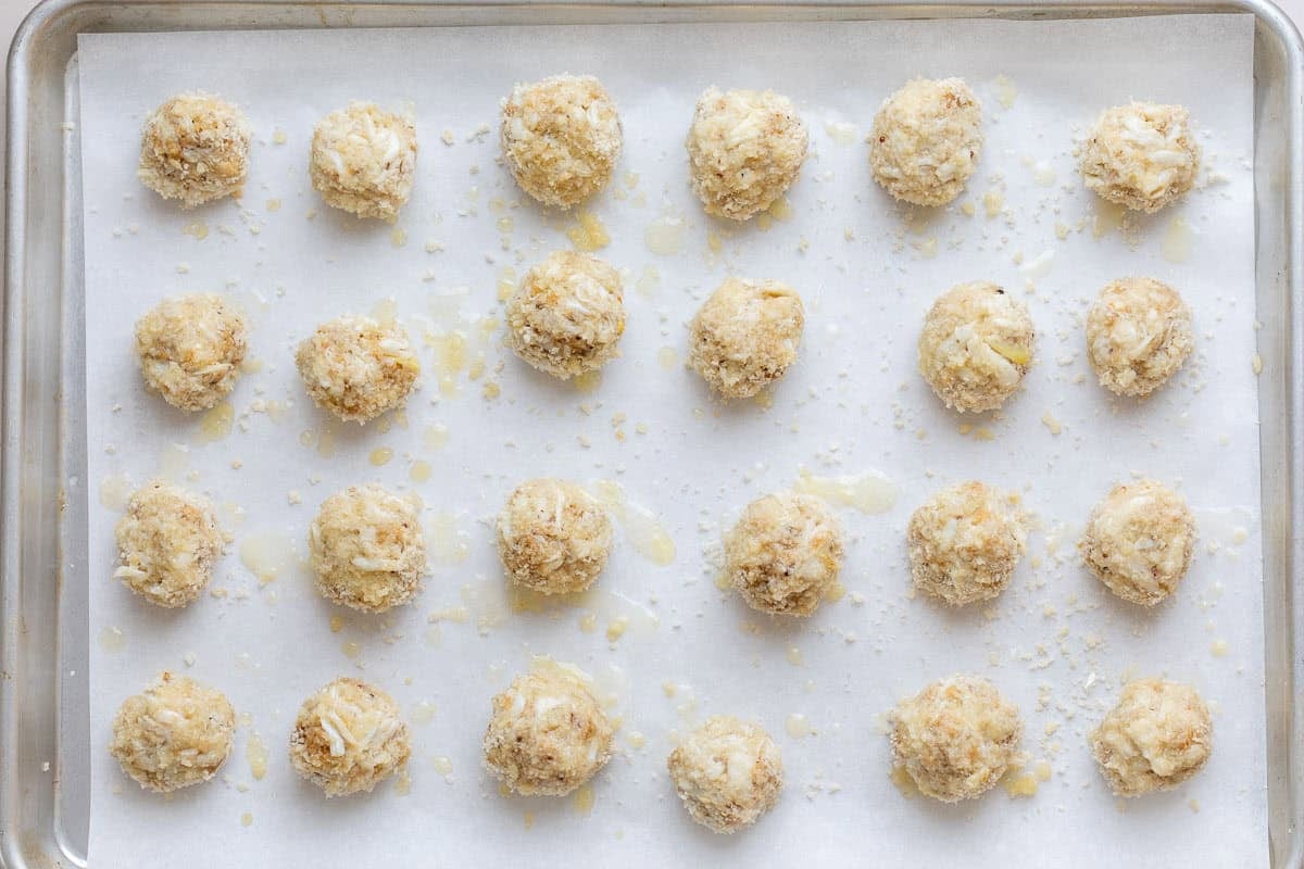 A baking tray lined with parchment paper holds evenly spaced, unbaked crab balls painted with butter ready for the oven.