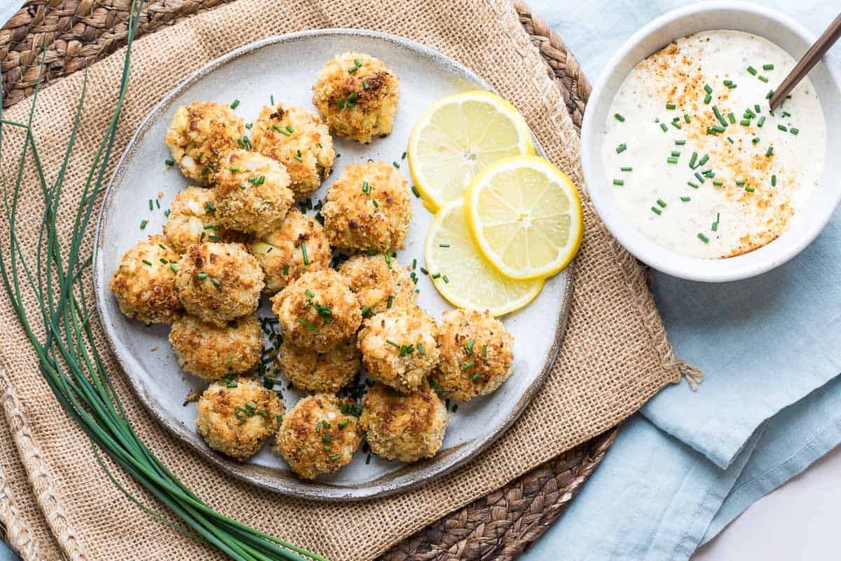 A plate of breaded, baked crab balls garnished with chives, lemon slices on the side, and a bowl of creamy dipping sauce with herbs.