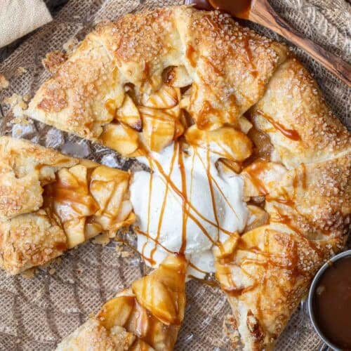 A rustic apple galette topped with a scoop of vanilla ice cream and drizzled with caramel sauce, served on a brown textured surface.