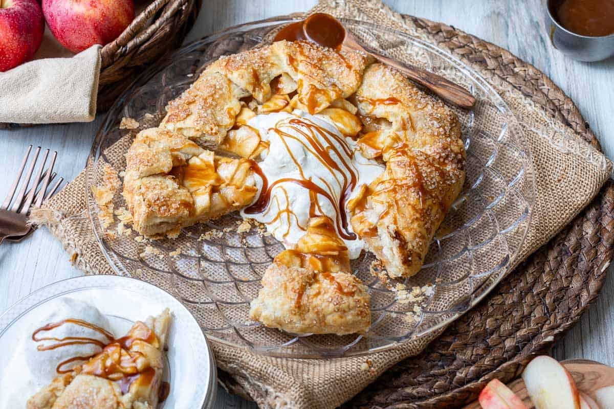 Rustic apple galette topped with ice cream and caramel sauce on a glass plate, with a slice served on a small plate nearby.