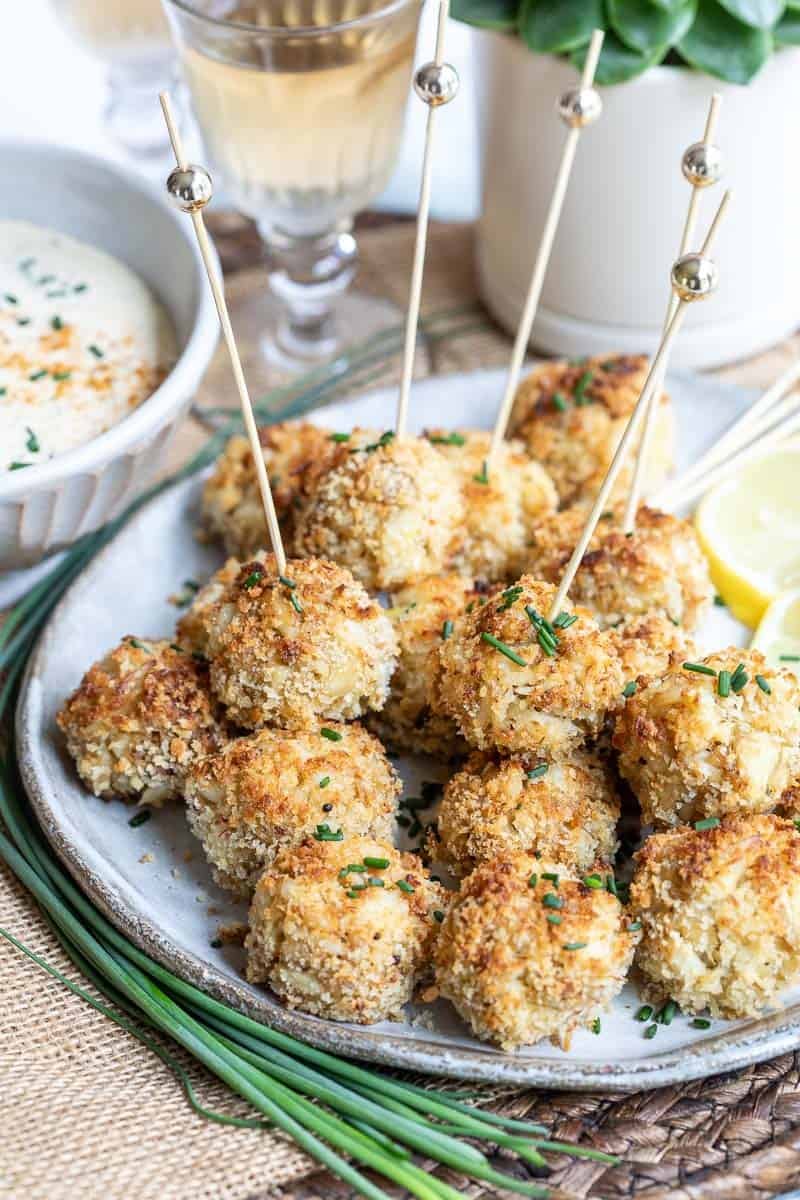 A plate of breaded, baked appetizer crab balls with wooden toothpicks, garnished with chopped chives, lemon slices, and sauce in the background.