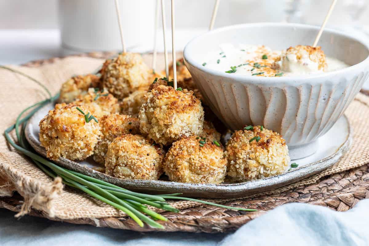 A plate of breaded, baked crab balls garnished with herbs, served with a bowl of creamy dipping sauce and wooden skewers on a woven placemat.