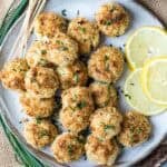 A plate of breaded, baked crab balls garnished with chopped chives, served with lemon slices and skewers on the side.