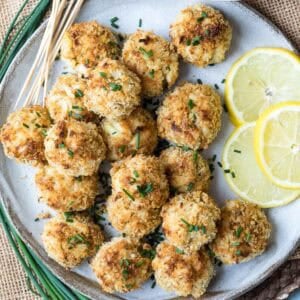 A plate of breaded, baked crab balls garnished with chopped chives, served with lemon slices and skewers on the side.