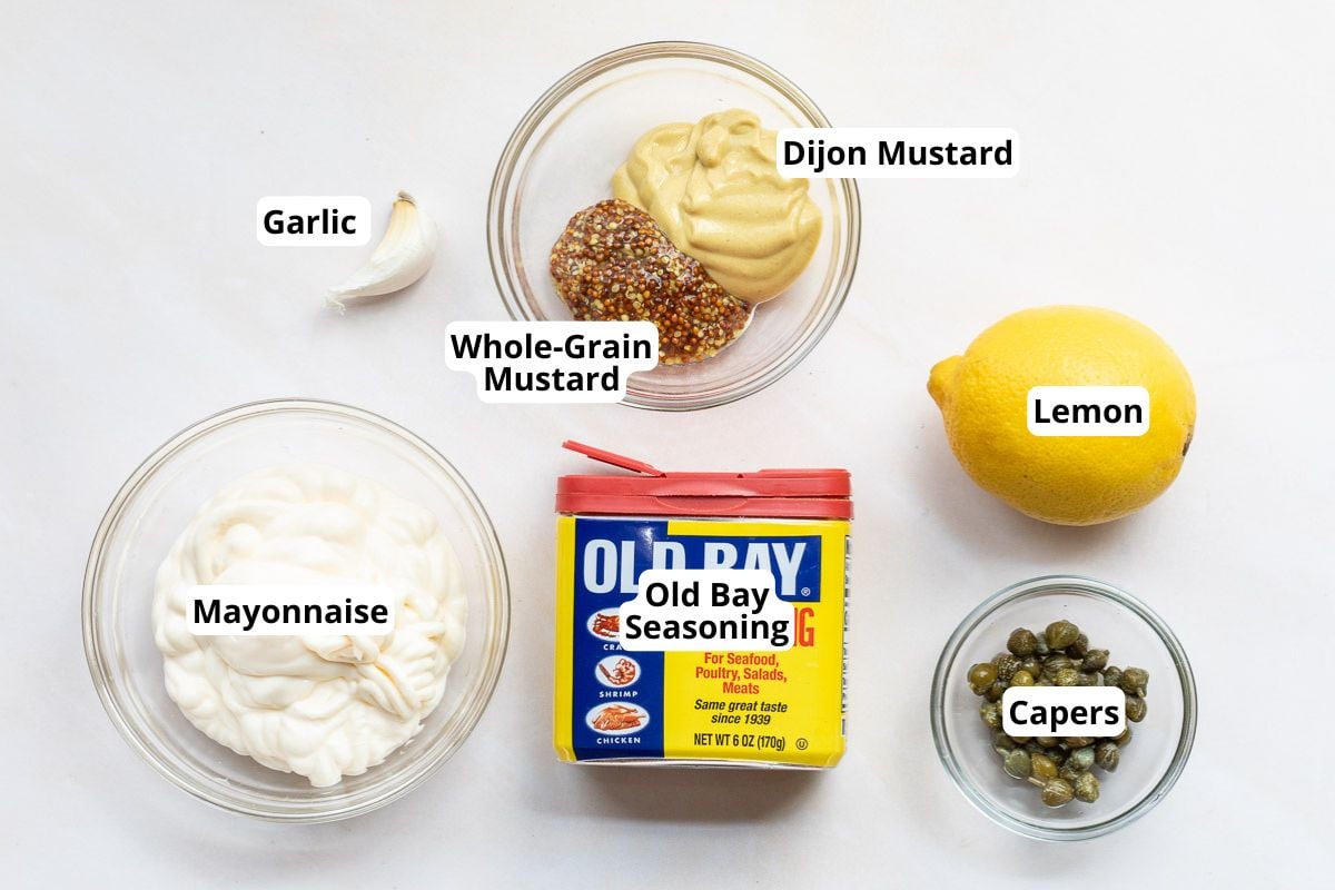 Assorted ingredients on a white surface: mayonnaise, garlic, whole-grain and Dijon mustard, lemon, Old Bay seasoning, and capers, each in a labeled bowl or container.