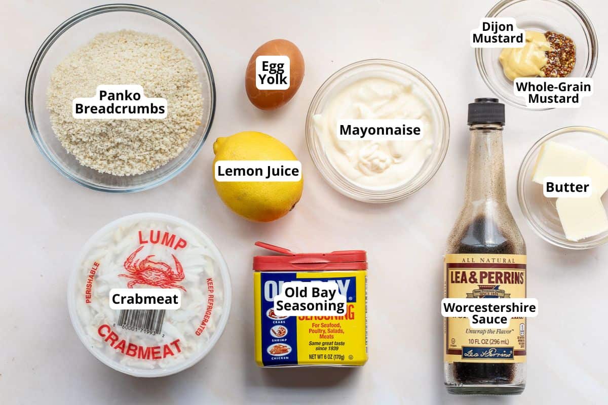Overhead view of labeled ingredients for crab cakes, including crabmeat, panko breadcrumbs, egg yolk, lemon juice, mayonnaise, mustards, butter, Old Bay seasoning, and Worcestershire sauce.