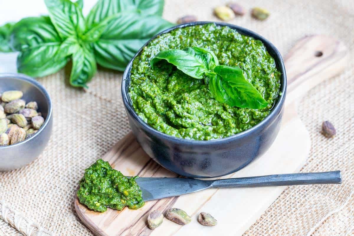 A bowl of pistachio pesto garnished with basil leaves, placed on a wooden board with a spreading knife and scattered pistachios nearby.