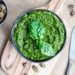 A bowl of pistachio pesto garnished with fresh basil leaves sits on a small wooden board, with pistachios nearby on a burlap surface.