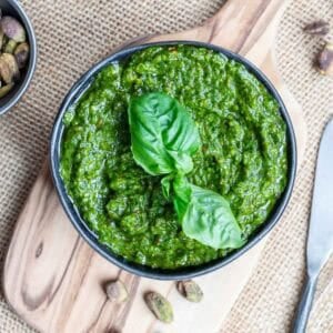 A bowl of pistachio pesto garnished with fresh basil leaves sits on a small wooden board, with pistachios nearby on a burlap surface.