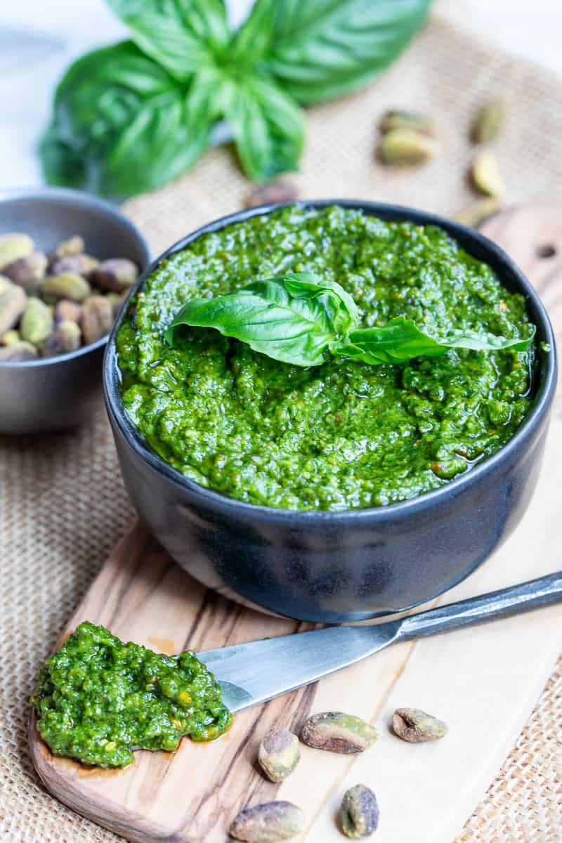 A bowl of pistachio pesto garnished with basil sits on a wooden board, with a knife, pistachios, and basil leaves nearby.