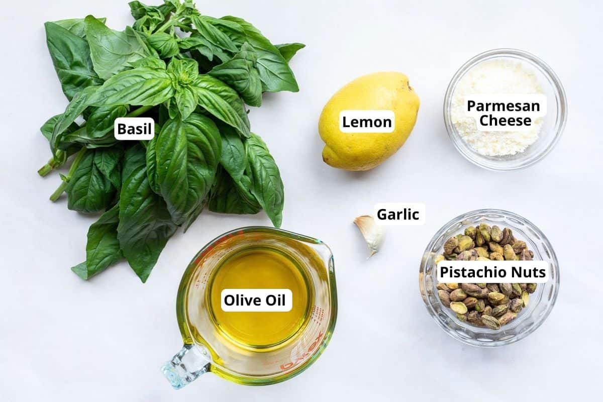 Ingredients for pesto include basil, lemon, parmesan cheese, olive oil, garlic, and pistachio nuts, each item labeled and displayed on a white background.