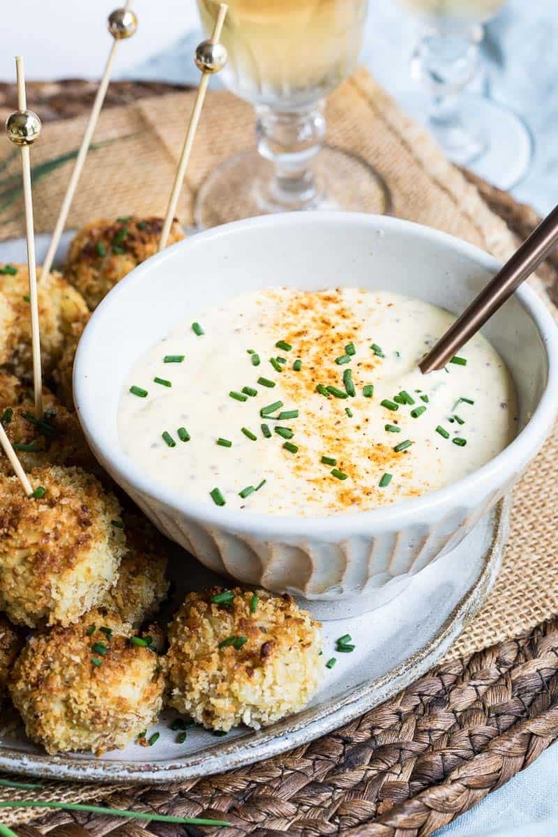 A bowl of creamy sauce for crab cakes garnished with chives and Old Bay Seasoning, surrounded by breaded bite-sized crab balls on skewers, set on a woven mat.