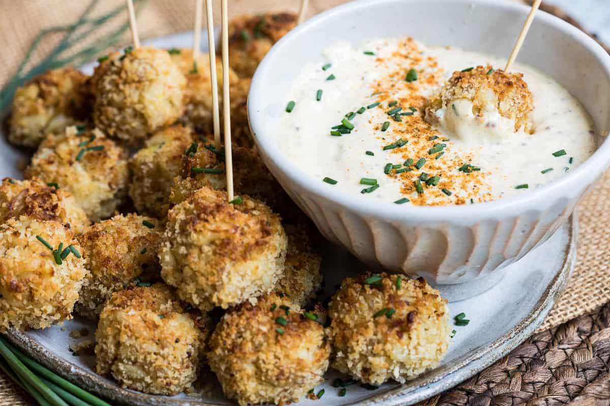 A plate of breaded, baked mini crab cakes with toothpicks, served with a bowl of creamy dipping sauce topped with herbs and spices.