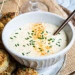White ceramic bowl filled with creamy dipping sauce for crab cakes, garnished with chopped chives and old bay seasoning, with a spoon resting inside. Baked crab balls are partially visible on the side.