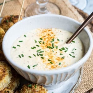 White ceramic bowl filled with creamy dipping sauce for crab cakes, garnished with chopped chives and old bay seasoning, with a spoon resting inside. Baked crab balls are partially visible on the side.