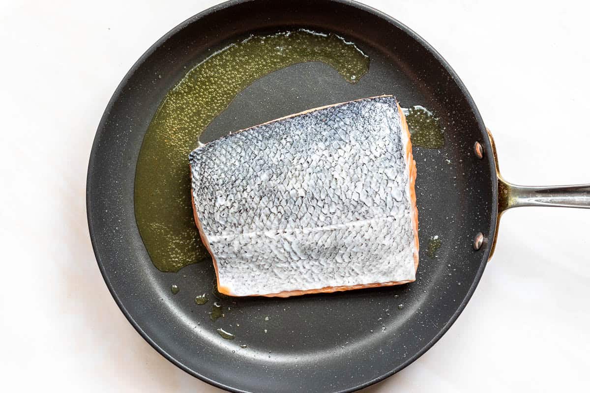 A skin-on salmon fillet searing flesh-side down in a frying pan with oil.
