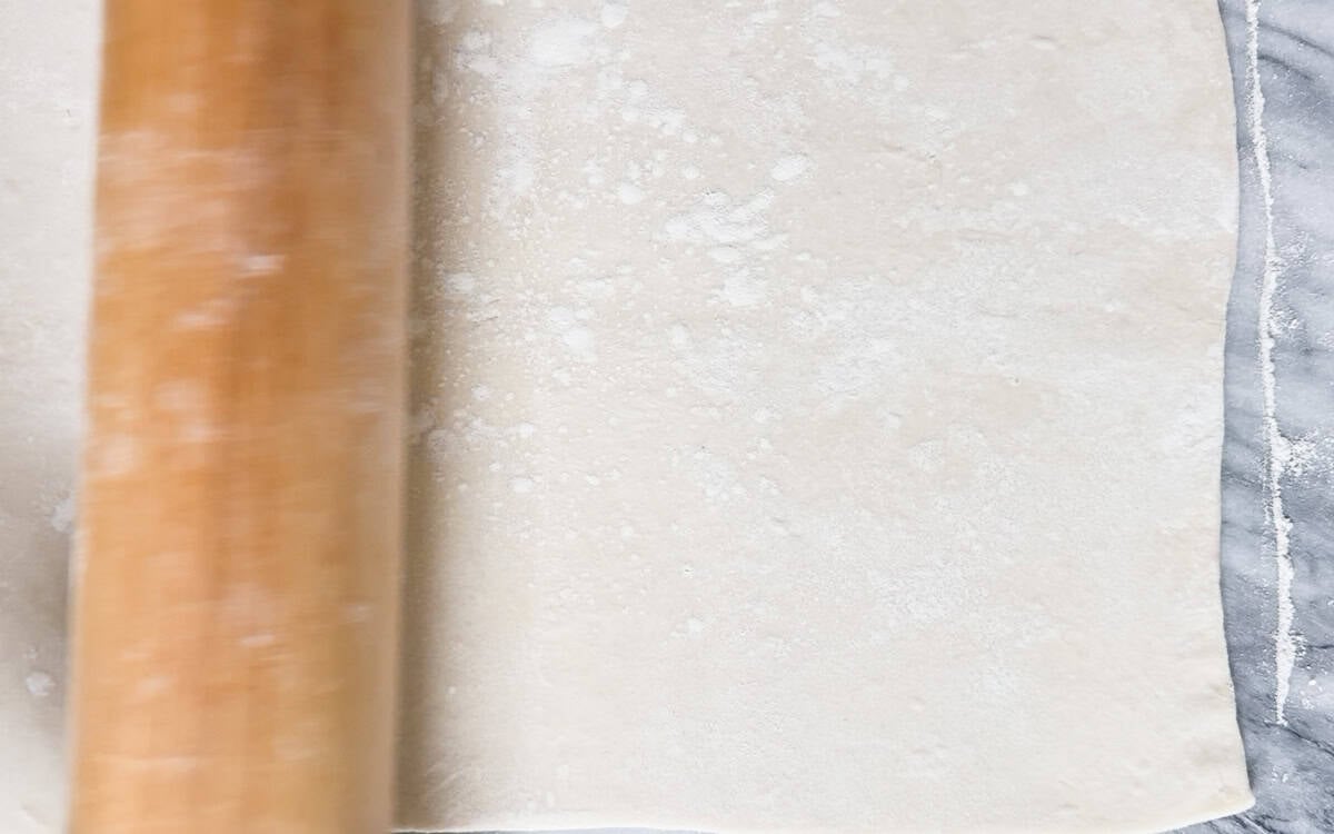 A wooden rolling pin flattening a sheet of dusted dough on a marble surface.