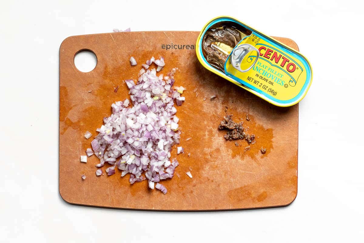 A pile of diced shallots and a small amount of minced anchovies on a wooden cutting board next to an open tin of Cento flat fillet anchovies.