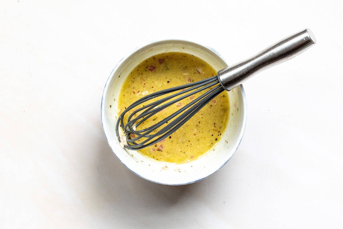 A wire whisk resting in a bowl containing a vinaigrette with shallots and pepper.