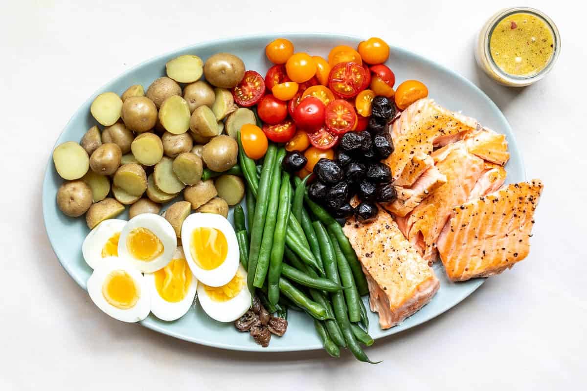 A platter of roasted potatoes, halved hard-boiled eggs, green beans, cherry tomatoes, black olives, and cooked salmon fillets, served with a side of dressing.