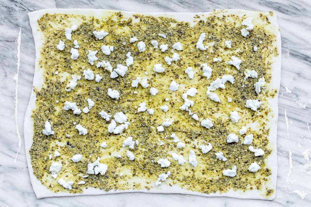 Puff pastry dough topped with pesto and goat cheese crumbles on a marble cutting board.