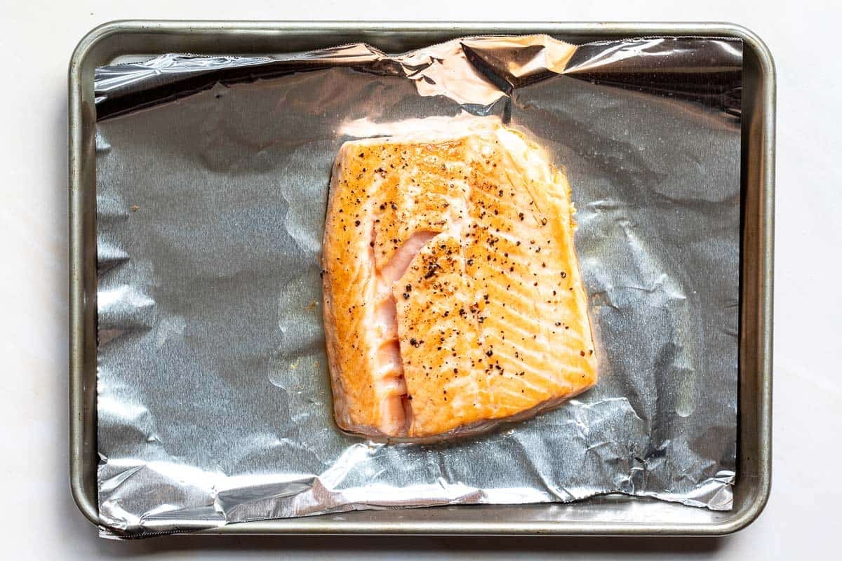 A seared and cooked salmon fillet seasoned with black pepper, resting on a foil-lined baking sheet.