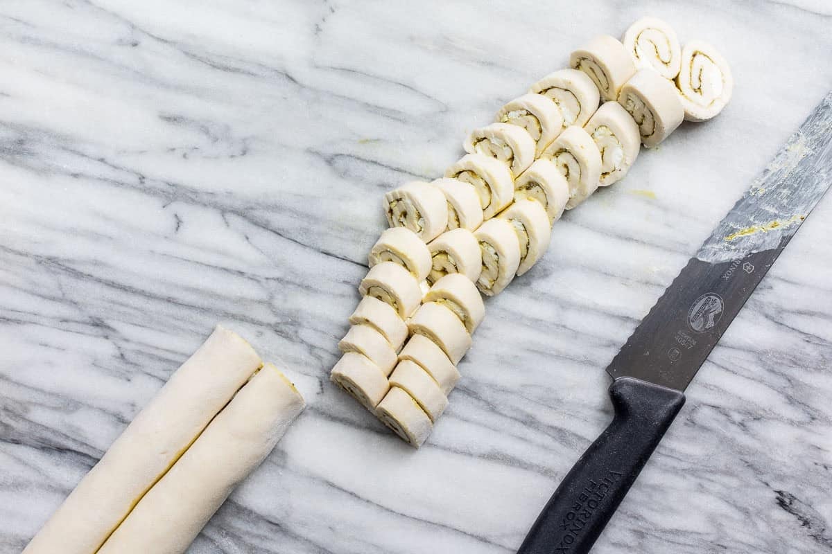 Sliced raw savory palmiers filled with pesto and goat cheese on a marble surface, a knife with some residue, and an unsliced roll are visible.
