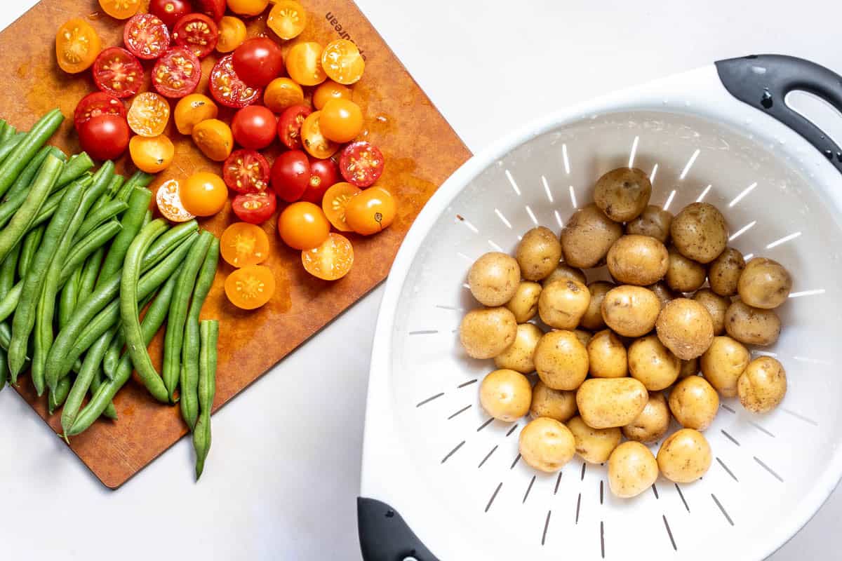 Green beans and halved cherry tomatoes on a wooden cutting board next to a white colander filled with small potatoes.