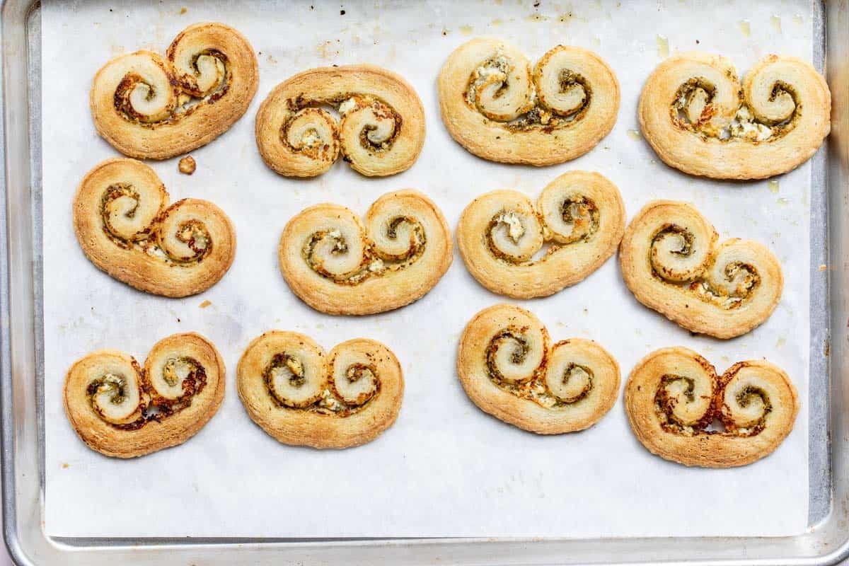 Twelve baked pesto palmiers with a savory filling are arranged on a parchment-lined baking sheet.