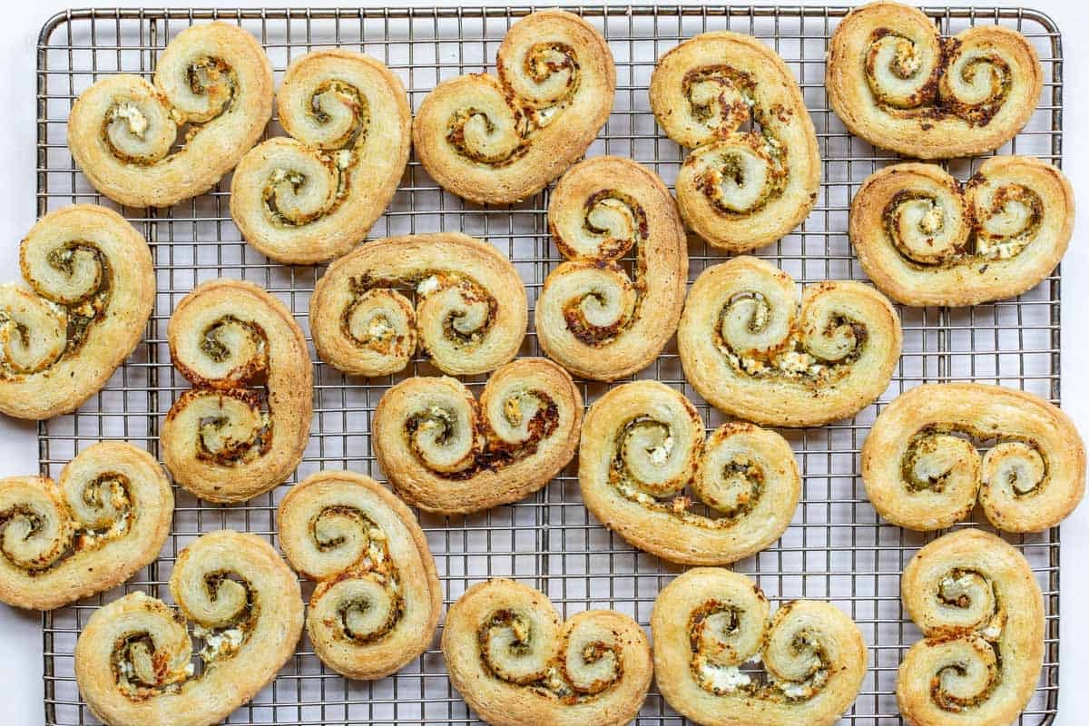 A cooling rack with 19 golden-brown savory palmiers with a visible pesto and goat cheese filling, arranged in a scattered pattern.
