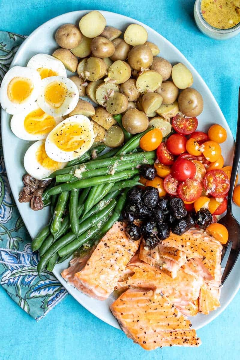 A platter of Salmon Niçoise-style salad with salmon fillets, halved soft-boiled eggs, green beans, halved cherry tomatoes, black olives, and roasted potatoes.