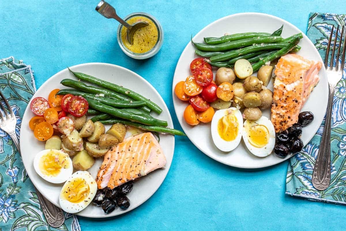 Two plates of salmon Niçoise salad with grilled salmon, hard-boiled eggs, green beans, halved cherry tomatoes, potatoes, and black olives, with a small jar of dressing.