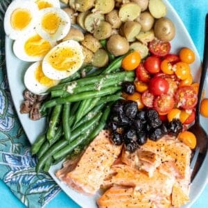 A platter of Salmon Niçoise salad with salmon, hard-boiled eggs, roasted potatoes, green beans, olives, and halved cherry tomatoes.