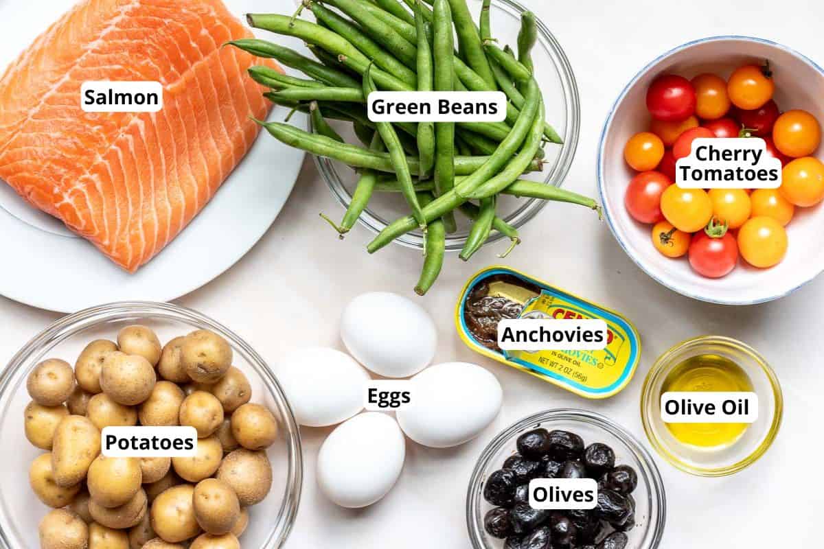Ingredients for a salmon Niçoise salad arranged on a surface: salmon fillet, green beans, cherry tomatoes, baby potatoes, eggs, canned anchovies, black olives, and a small bowl of olive oil.