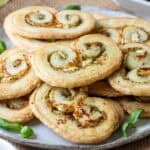 A plate of golden, flaky puff pastry palmiers filled with green pesto and cheese, garnished with fresh basil leaves.