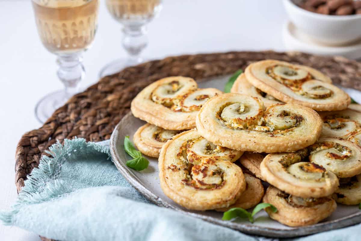 A plate of spiral-shaped puff pastry palmiers sits on a woven placemat, with two glasses of white wine and a bowl of nuts in the background.