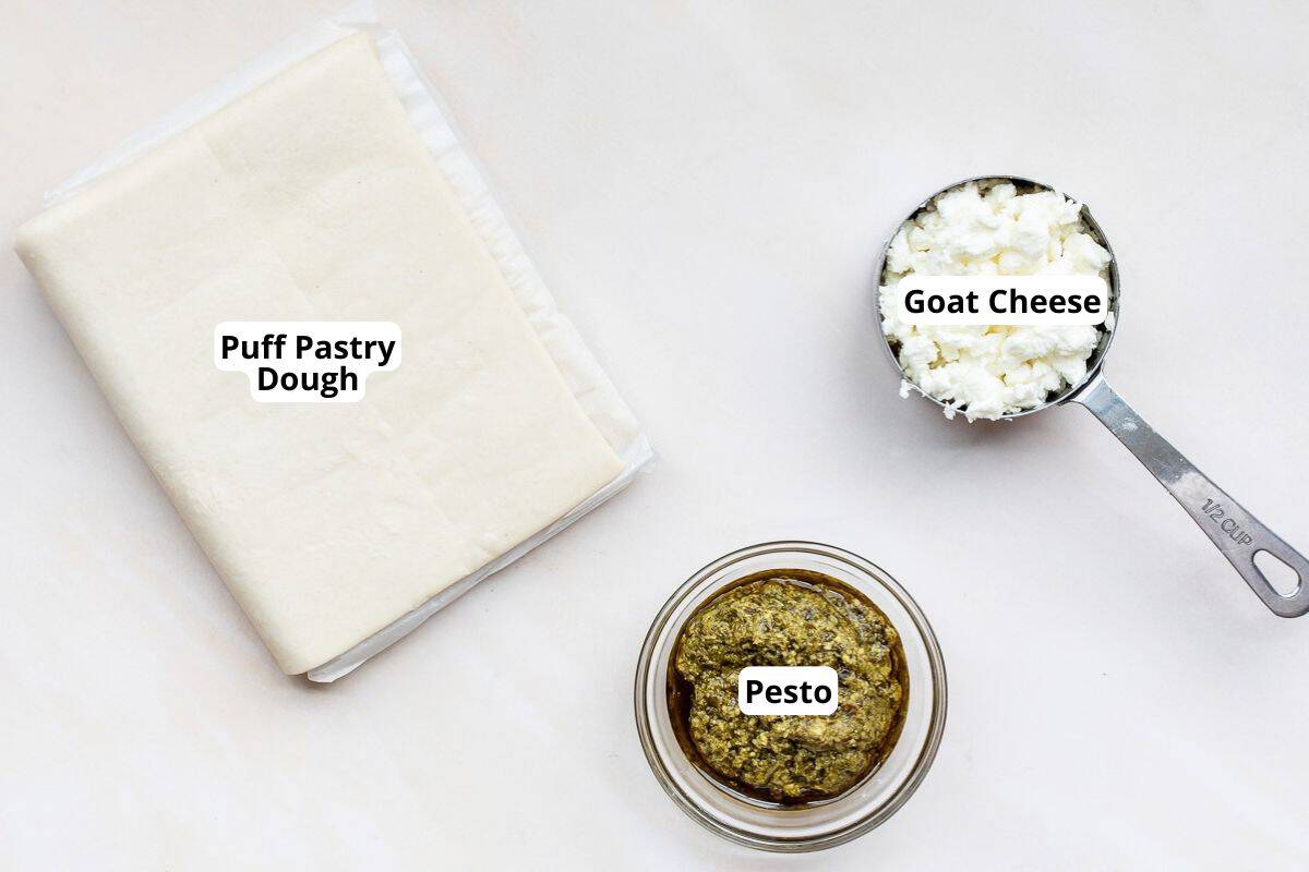 Ingredients for pesto palmiers: a folded sheet of puff pastry dough, a half-cup measuring cup filled with crumbled goat cheese, and a small glass bowl of pesto.